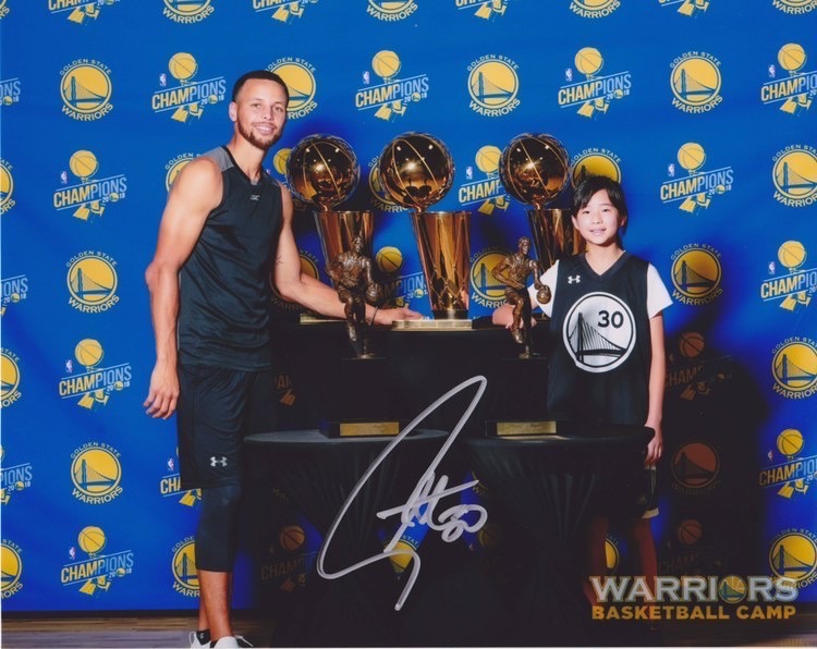 Tang poses for a photo with Curry at Warriors Basketball Camp surrounded by various championship and MVP trophies.