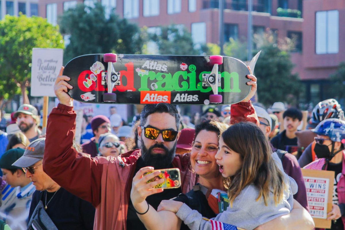 A family of three poses for a selfie as they walk across Lefty O’Doul Bridge, displaying a skateboard decorated in Black History Month colors with the slogan “end racism.” Photo | Isabelle Kok