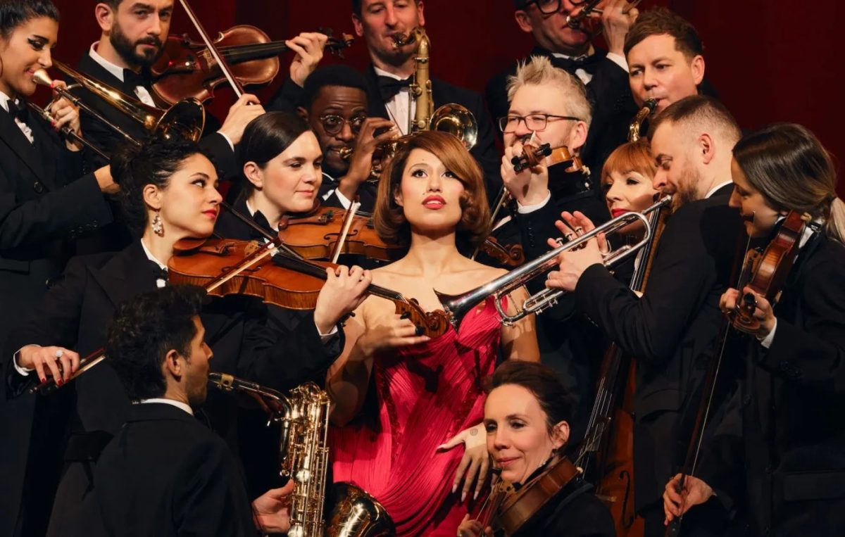 RAYE poses in vibrant red dress while surrounded by an orchestra, a nod to both her flashy style and theatric, orchestral music. Photo | Aliyah Otchere